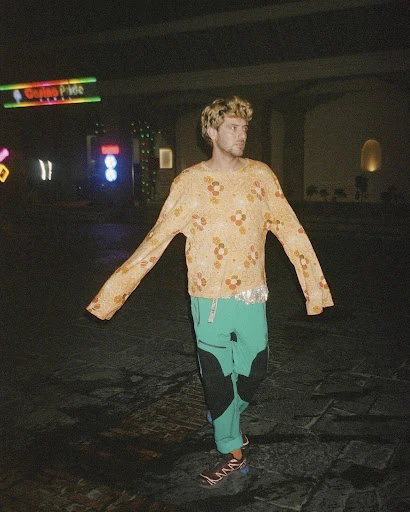 Adult male poses with arms outstretched in patterned shirt and pants in outdoor urban environment at night with neon lights.