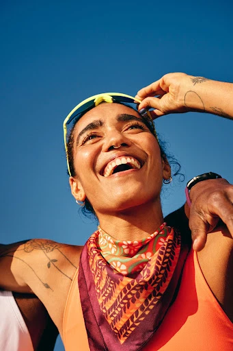 Smiling woman with a colorful scarf and sunglasses on her head gazes upwards under a clear blue sky in a bright, sunny outdoor setting.