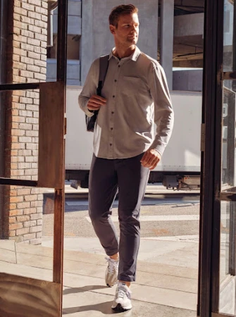 Man walking through an open glass door wearing a light-coloured long-sleeve shirt, dark trousers, and white sneakers.