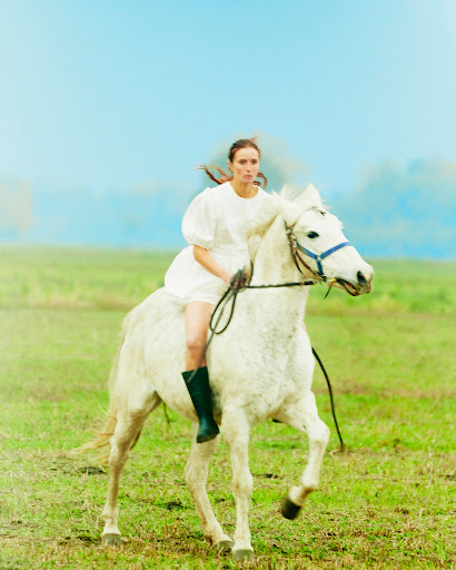 Woman in a white dress riding a white horse across a grassy field under a pale sky