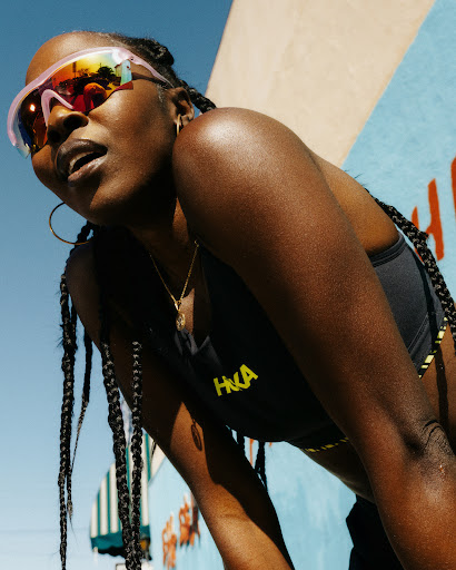 Woman with braids wears athletic sunglasses and workout gear near a colorful mural outdoors on a sunny day with a low angle.