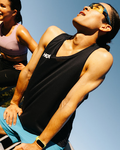 Two athletes take a break outdoors with the man in the foreground wearing a black athletic top and blue shorts, the woman is in the background.