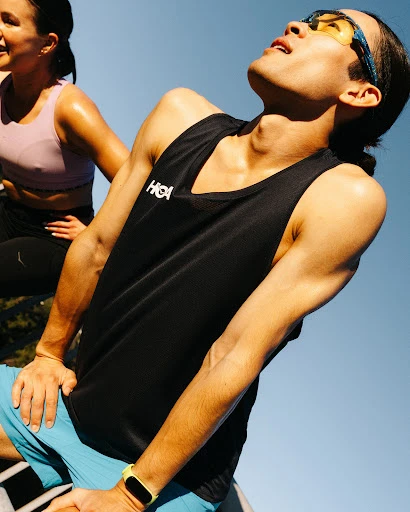 Two athletes take a break outdoors with the man in the foreground wearing a black athletic top and blue shorts, the woman is in the background.