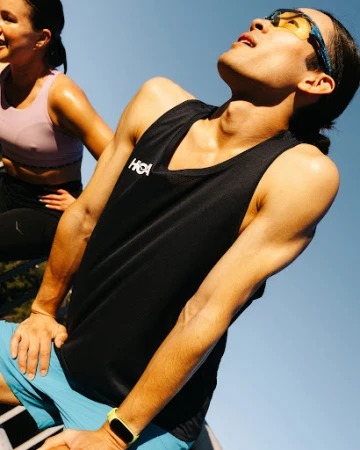 Two athletes take a break outdoors with the man in the foreground wearing a black athletic top and blue shorts, the woman is in the background.