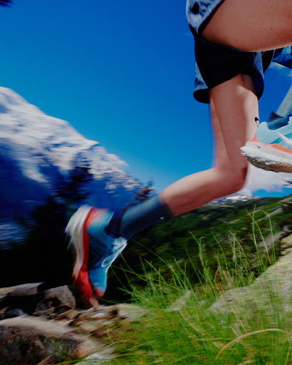 Woman running on a rocky trail with mountain background, wearing athletic shoes, shorts, and socks, captured in motion with blurred background.