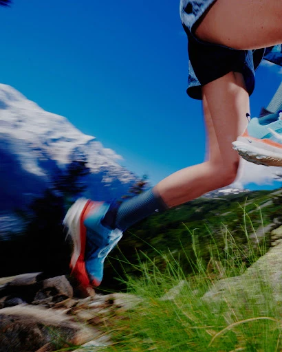 Woman running on a rocky trail with mountain background, wearing athletic shoes, shorts, and socks, captured in motion with blurred background.