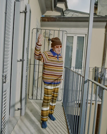 Elderly adult waves from balcony, wearing patterned knitwear, hat, and heart-shaped glasses, set against a neutral-toned building exterior.