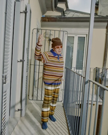 Elderly adult waves from balcony, wearing patterned knitwear, hat, and heart-shaped glasses, set against a neutral-toned building exterior.