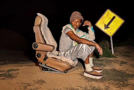 Person sitting on chair next to a road sign in an outdoor setting. They are wearing casual clothing and shoes with platform soles.