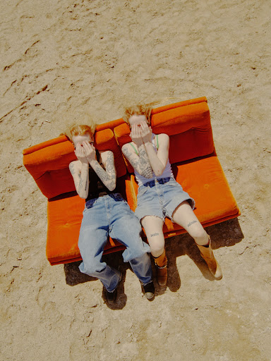Two women pose on an orange couch in a desert setting, covering their faces with their hands. Both wear denim shorts or pants.