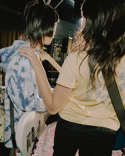 Two musicians standing in a music studio, one with a bass guitar and the other wearing a t-shirt with floral patterns on the back.