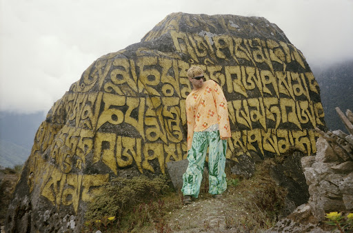 Blond adult wearing sunglasses, patterned orange top and green pants standing next to a large rock with inscribed yellow characters on it.