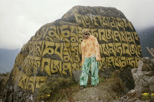 Blond adult wearing sunglasses, patterned orange top and green pants standing next to a large rock with inscribed yellow characters on it.