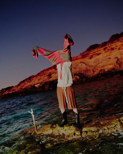 A fashion shot on a rocky shore at dusk. A person wears a skirt, boots, and is holding a pink patterned garment overhead. Waves crash.