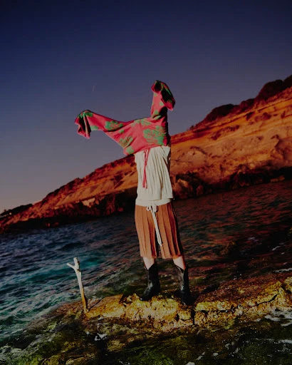 A fashion shot on a rocky shore at dusk. A person wears a skirt, boots, and is holding a pink patterned garment overhead. Waves crash.