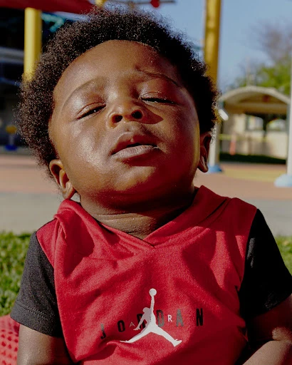 Close-up of a baby with dark skin, wearing a red Jordan hooded shirt, eyes closed, outdoors on a sunny day in a residential area.
