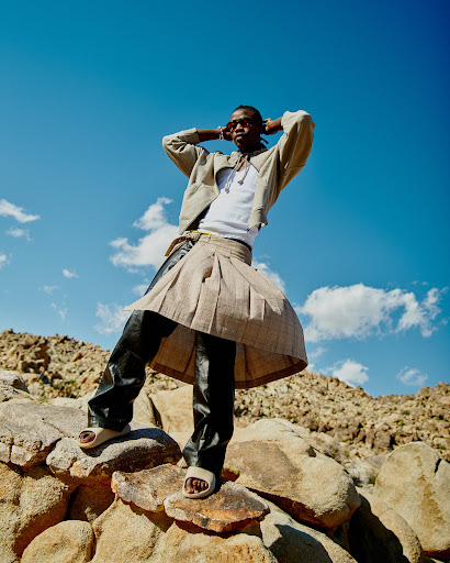 Young Black man poses on desert rocks wearing jacket, skirt, and sandals against blue sky with clouds. Fashion editorial photography.