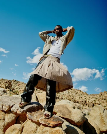 Man standing on rocky terrain wearing a pleated wrap skirt, jacket, and sunglasses against a clear blue sky.