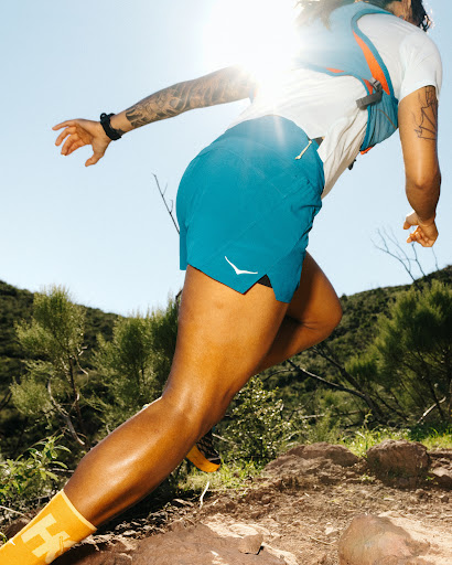 Adult runs uphill on a rocky trail in turquoise shorts, white t-shirt, and yellow socks in bright daylight, green trees in the background.
