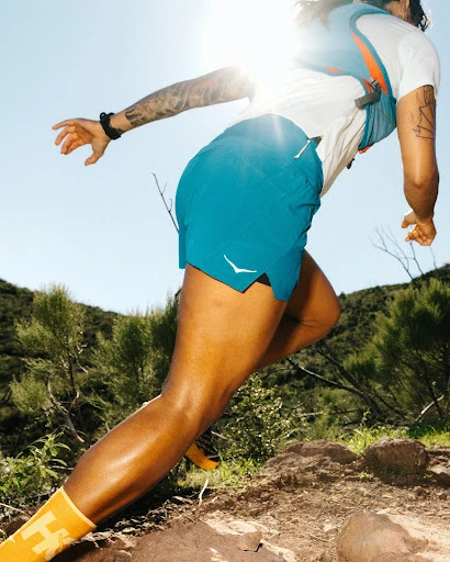 Adult runs uphill on a rocky trail in turquoise shorts, white t-shirt, and yellow socks in bright daylight, green trees in the background.