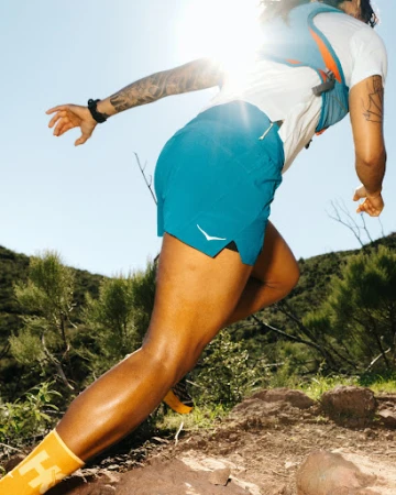 Adult runs uphill on a rocky trail in turquoise shorts, white t-shirt, and yellow socks in bright daylight, green trees in the background.