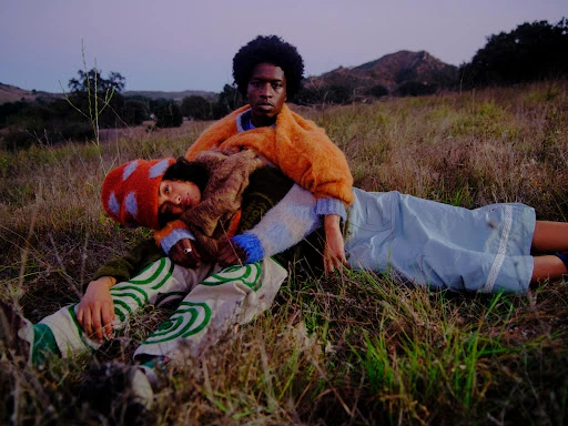 Two models resting on a grassy meadow at dusk wearing textured knitwear and patterned trousers styled by Charlotte Patterson