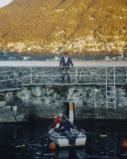 Two men in a rowboat on a lake with buildings on a distant hillside, viewed from a stone pier with a man standing above.