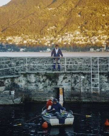 Two men in a rowboat on a lake with buildings on a distant hillside, viewed from a stone pier with a man standing above.