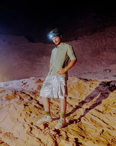 Adult male poses on desert rock formation wearing headlamp, short-sleeve shirt, shiny shorts, and sneakers in dimly lit cave interior.