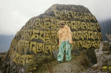 Man standing in front of a massive boulder covered in carved yellow Tibetan script, wearing patterned orange top and green trousers.