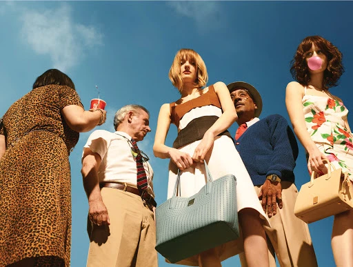 Fashion editorial featuring diverse individuals against a bright blue sky. One woman blows a pink bubble with gum.