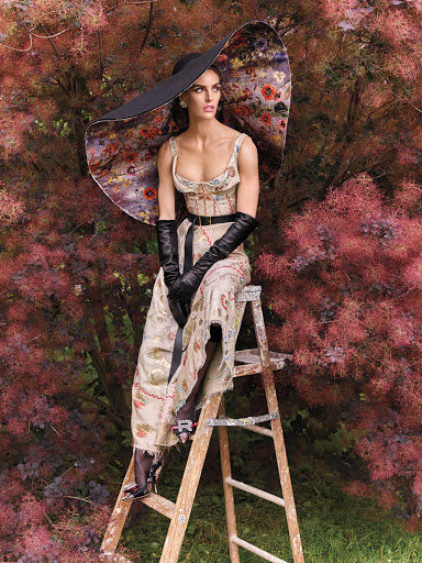 A woman in a floral dress and large hat sits on a ladder surrounded by trees with red foliage in an outdoor fashion shot.