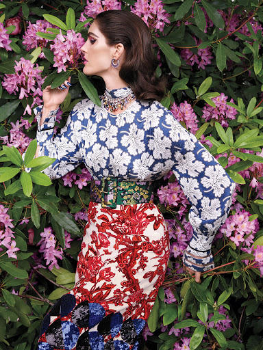 Adult female fashion model poses with floral patterned shirt and skirt, jewelry in natural outdoor background of green leaves and pink flowers.