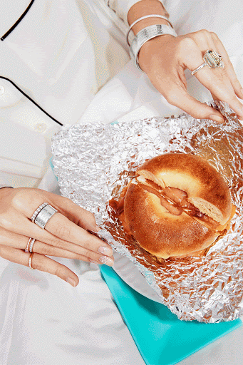 Close-up shot of adult hands holding a bagel sandwich wrapped in foil, showcasing rings and bracelets against a white and turquoise backdrop.