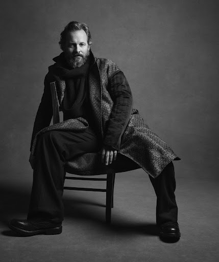 Black and white portrait of a bearded man sitting on a chair, wearing a patterned coat and dark clothing against a grey backdrop.