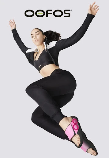 Athletic woman in black sportswear jumps in the air, wearing pink and black OOFOS recovery sandals against a grey backdrop with the brand logo.