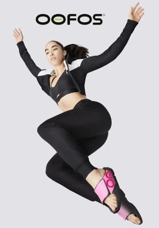 Athletic woman in black sportswear jumps in the air, wearing pink and black OOFOS recovery sandals against a grey backdrop with the brand logo.