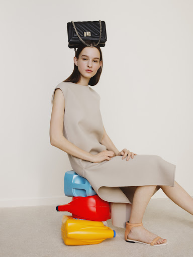 Woman poses indoors wearing a dress with a handbag on her head, sitting on colorful plastic containers against a neutral backdrop.