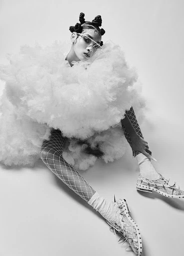 Monochrome fashion shot of a person with creative hair and makeup wearing a puffy white dress, fishnet stockings, and embellished shoes.