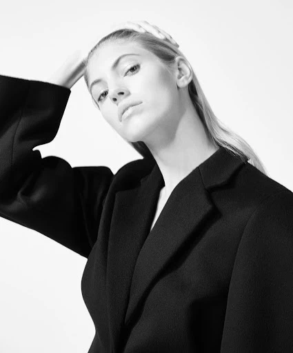Black and white portrait of a young woman in a dark blazer against a bright background, hands near her head, looking at camera.