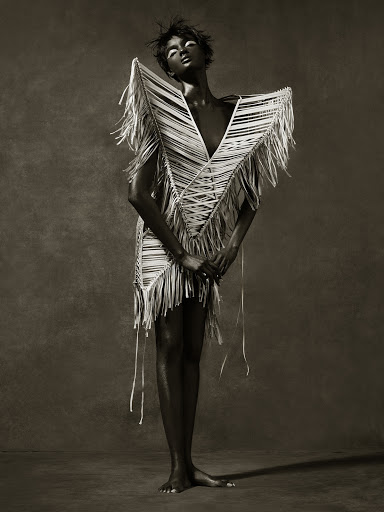 Fashion shot of an African American woman in a sculptural fringed dress, striking a pose against a textured gray background in studio lighting.