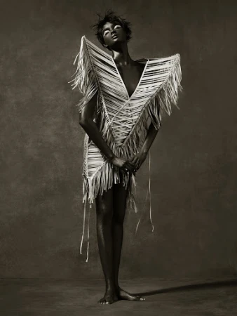 Model in avant-garde fringe dress with dramatic white eye makeup against a textured dark background