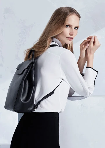Fashion shot of a woman in a white blouse and black skirt with a black leather backpack, posing against a light patterned background.