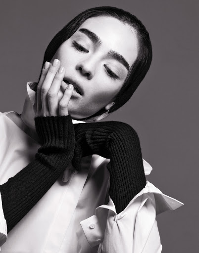 A young adult woman with dark hair, wearing a white collared shirt and black knit arm warmers, poses in a black and white portrait.