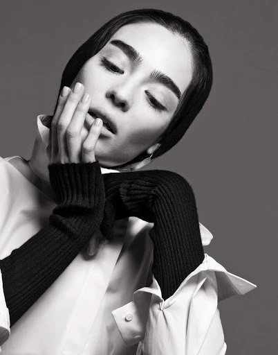 A young adult woman with dark hair, wearing a white collared shirt and black knit arm warmers, poses in a black and white portrait.
