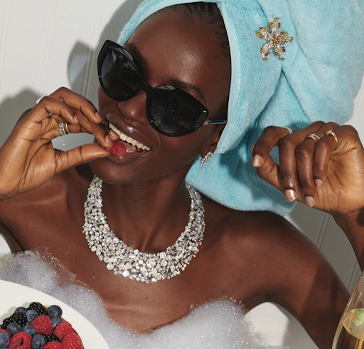 An adult Black woman enjoys berries in a bubble bath, wearing sunglasses, jewelry, and a hair towel with decorative flower clip.