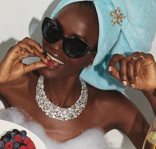 An adult Black woman enjoys berries in a bubble bath, wearing sunglasses, jewelry, and a hair towel with decorative flower clip.