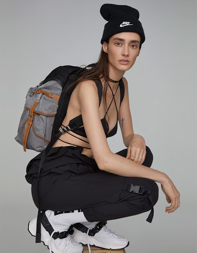 Model wearing black beanie, black strappy top, cargo pants, and white sneakers, crouching against grey seamless studio background.