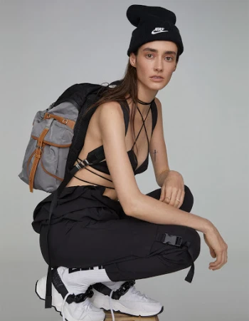 Model wearing black beanie, black strappy top, cargo pants, and white sneakers, crouching against grey seamless studio background.