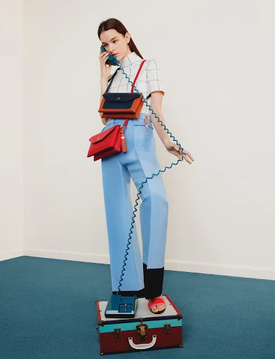 A woman in blue pants and a grid shirt stands on a suitcase while talking on a vintage telephone with two crossbody bags.